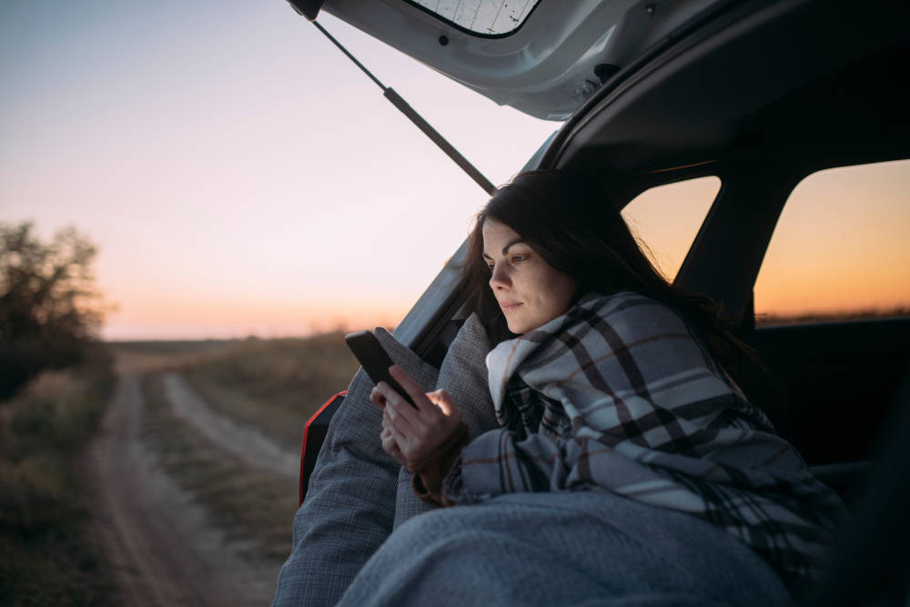 En tjej sitter i en bil och tittar på en mobil i solnedgången.